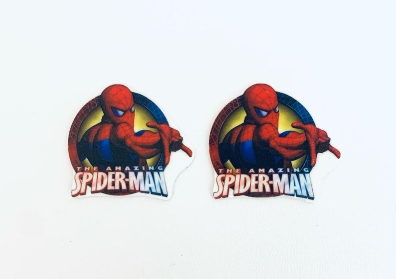 以下が含まれることがあります： アメージング・スパイダーマンのロゴが描かれた、赤、青、黄色の丸いステッカー2枚。ロゴには、赤いスーツと青いスーツを着たスパイダーマンの漫画が描かれており、ウェブデザインが施されています。テキスト「The Amazing Spider-Man」は白い文字で書かれています。
