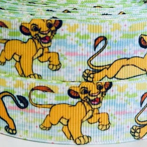 May include: A white ribbon with a repeating pattern of a cartoon lion cub in various poses. The lion cubs are yellow with black eyes and noses. The background is a light blue and green striped pattern with white dots.
