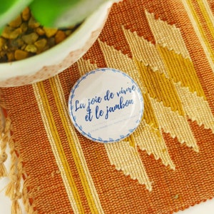 Peut inclure: Un bouton blanc avec du texte bleu qui dit "La joie de vivre et le jambon" sur un fond tissé orange et crème.