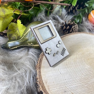 Può includere: Un ritaglio di legno di una console per videogiochi portatile vintage. La console è di colore marrone chiaro con uno schermo grigio e pulsanti neri. La console è appoggiata su una superficie di legno.