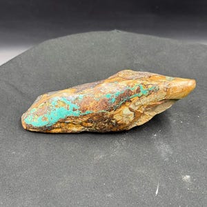 May include: A natural stone with a mix of colors, including turquoise, brown, and tan. The stone has a rough texture and irregular shape. The turquoise color is concentrated in patches, creating a striking contrast against the other earthy tones. This stone could be used for jewelry or decorative purposes.