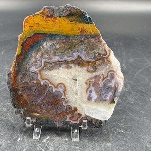 May include: A polished agate stone slice with a variety of colors, including yellow, orange, red, brown, and white. The stone is displayed on a clear acrylic stand, showcasing its natural patterns and textures. This decorative piece is ideal for home decor or as a unique gift.