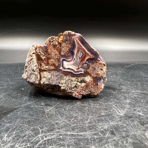 May include: A large, irregularly shaped agate geode with a natural, rough exterior. The interior of the geode is filled with a beautiful, swirling pattern of purple, white, and brown agate.