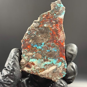 May include: A multi-coloured stone slab with a rough, uneven surface. The stone displays a dark brown base with streaks of red, turquoise, and grey. The stone is held by a gloved hand.
