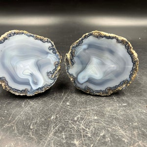 May include: Two agate slices with a white and gray swirling pattern. The edges of the slices are a dark gray color.