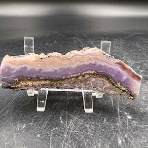 May include: A polished slab of purple agate with brown and white banding. The agate is displayed on a clear acrylic stand.
