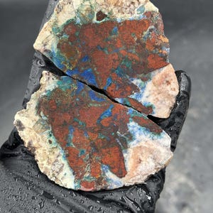 Puede incluir: Dos piezas de una piedra cortada con un patrón llamativo. La piedra presenta una mezcla de colores, incluyendo rojo intenso, azul y verde, sobre un fondo beige y blanquecino. La piedra es sostenida por una mano enguantada.