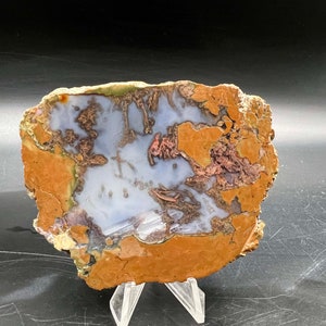 May include: A slice of agate with a white and light blue center, surrounded by a brown, orange, and green outer layer. The agate is on a clear stand.
