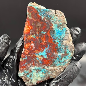 May include: A vibrant, raw gemstone slice with a striking mix of colors. The stone features a rich, rusty red base, contrasted by patches of bright turquoise and hints of gray. The natural, rough edges add to its unique appeal.