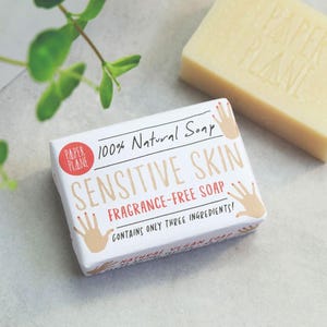 Puede incluir: Barra de jabón blanca con las palabras "Paper Plane" y "100% Natural Soap" impresas en ella. El jabón está etiquetado como "Sensitive Skin Fragrance-Free Soap" y dice "¡Contiene solo tres ingredientes!"