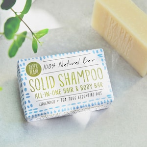 May include: Two bars of soap are shown, one wrapped and one unwrapped. The wrapped bar has text reading "SOLID SHAMPOO ALL-IN-ONE HAIR & BODY BAR". The unwrapped bar is a light yellow colour. The bars are for hair and body.