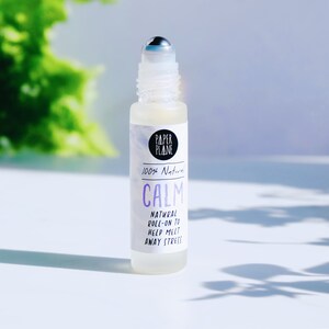 May include: A clear glass roller bottle with a silver roller ball. The bottle is labeled "PAPER PLANE" and "100% Natural CALM NATURAL ROLL-ON TO HELP MELT AWAY STRESS".