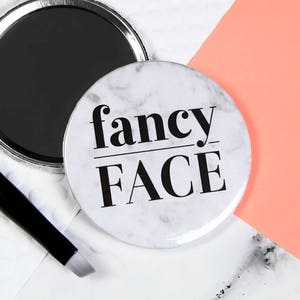 Puede incluir: Un espejo compacto blanco con un patrón de mármol y texto negro que dice "fancy FACE".