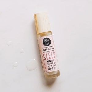 Könnte beinhalten: Eine Glas-Roll-on-Flasche mit hellem Holzdeckel. Das rosa Etikett trägt die Aufschrift "PAPER PLANE", "100% Natural" und "SLEEP". Der Text "NATURAL ROLL-ON TO HELP YOU DRIFT OFF" steht ebenfalls auf dem Etikett. Mehrere klare Flüssigkeitstropfen sind verstreut.