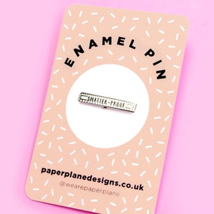 May include: A white enamel pin featuring a silver ruler design that reads "Shatter-Proof". The pin is displayed on a pink and white background with the text "enamel pin" and "paperplanedesigns.co.uk" in black.