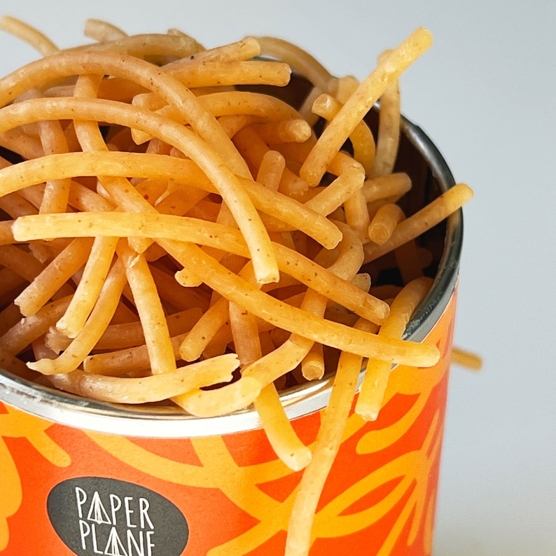 May include: A close-up of a can filled with thin, dry, golden brown noodles. The can is orange and has the words "Paper Plane" printed on it.