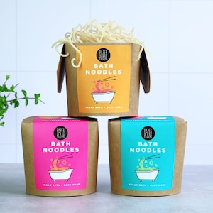 May include: Three cardboard boxes with a yellow, pink, and blue label. Each box has a graphic of a bowl of noodles and the text "Paper Plane Bath Noodles Vegan Bath + Body Wash".
