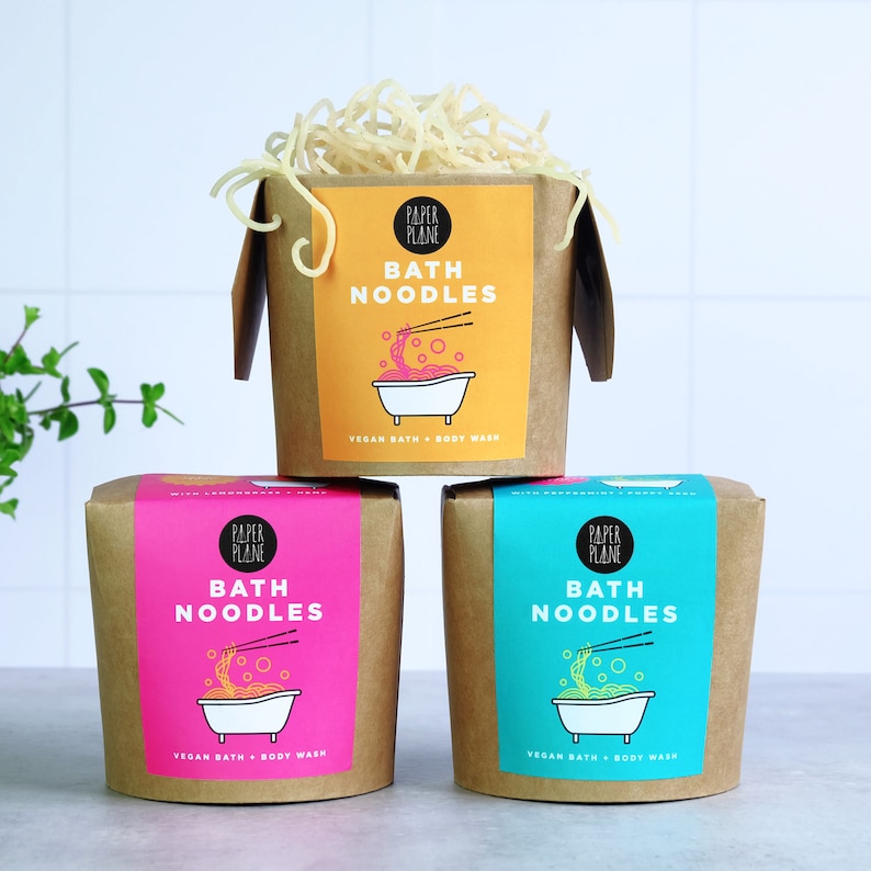 May include: Three cardboard boxes with a yellow, pink, and blue label. Each box has a graphic of a bowl of noodles and the text "Paper Plane Bath Noodles Vegan Bath + Body Wash".