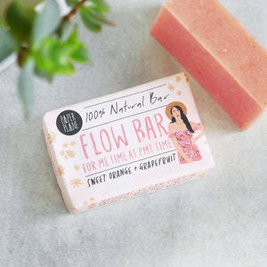 Pode incluir: Uma barra de sabonete rosa com um rótulo branco que diz "100% Natural Bar Flow Bar For Me Time at PMT Time Sweet Orange + Grapefruit". O rótulo apresenta uma ilustração de desenho animado de uma mulher vestindo um vestido floral e um chapéu de palha.