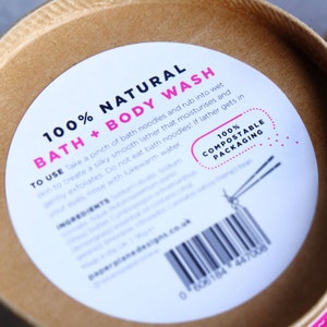 May include: A white label with pink and black text that reads "100% Natural Bath + Body Wash". The label also includes instructions for use, a list of ingredients, and a barcode. The label is on a brown cardboard tube.