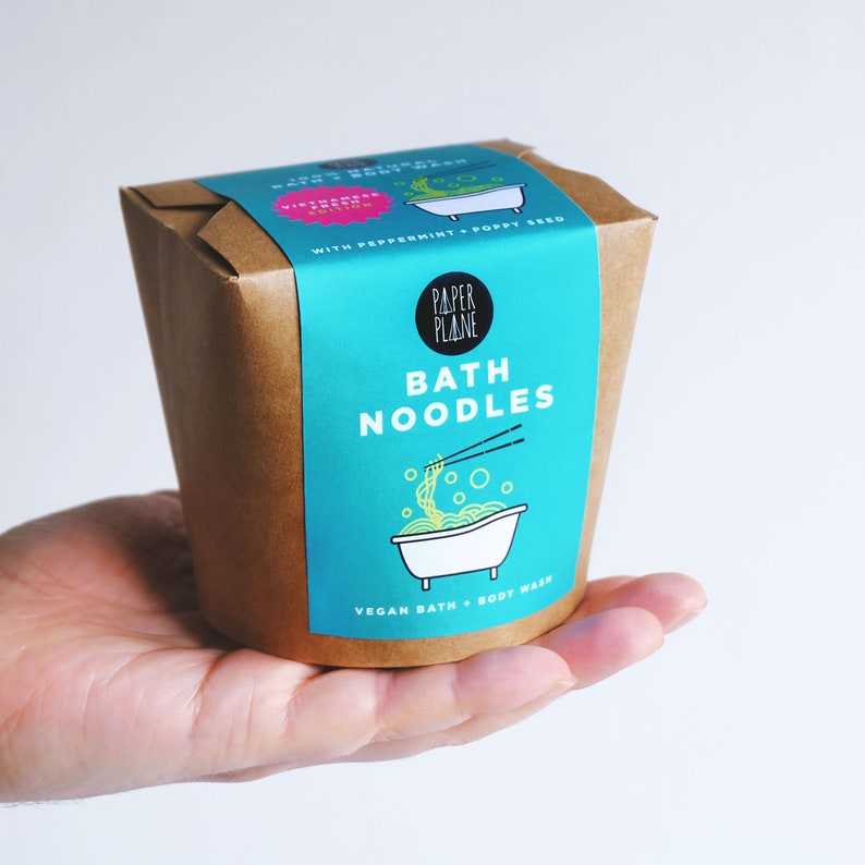 May include: A brown paper takeout container with a blue and white label that says "Bath Noodles" and "Vegan Bath + Body Wash". The label features a cartoon illustration of a bathtub with noodles and chopsticks.