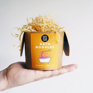 May include: A brown paper takeout container filled with yellow bath noodles. The container has a label that says "Paper Plane Bath Noodles Vegan Bath + Body Wash".
