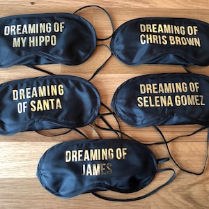 Puede incluir: Cinco antifaces negros con texto dorado. El texto en las antifaces dice "Dreaming of" seguido de un nombre. Los nombres son "My Hippo", "Chris Brown", "Santa", "James" y "Selena Gomez".