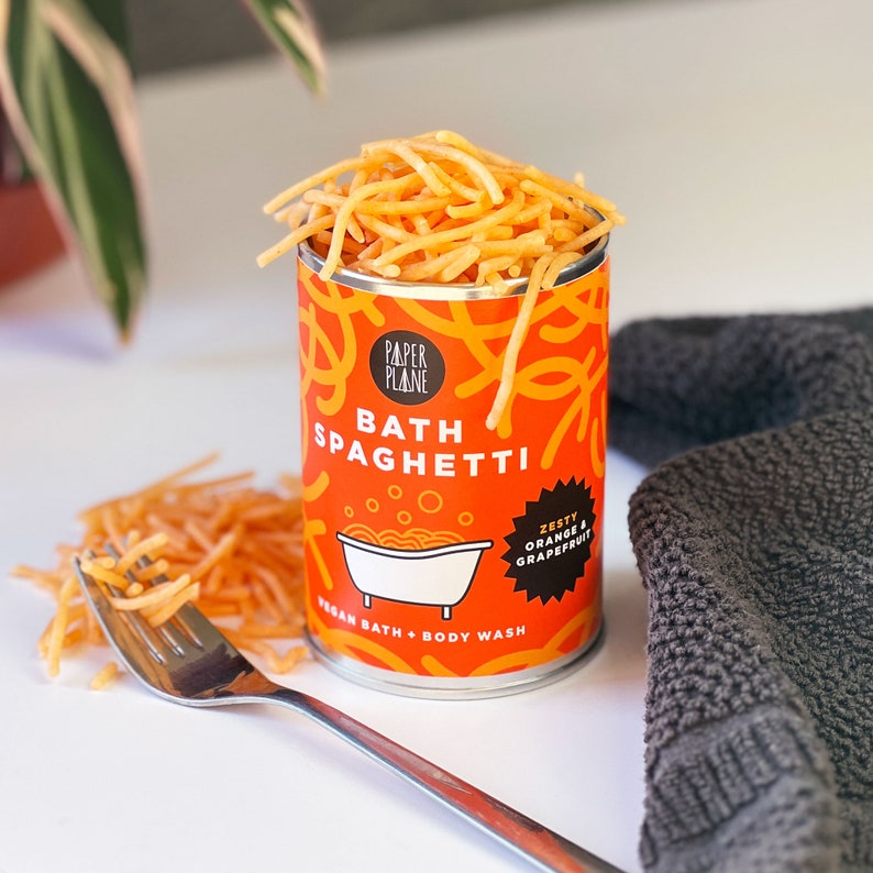 May include: A can of orange and grapefruit scented bath spaghetti, a vegan bath and body wash, with the text "Paper Plane Bath Spaghetti Zesty Orange & Grapefruit Vegan Bath + Body Wash" on the label.