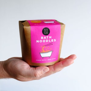 May include: A brown paper container with a pink label that says "Bath Noodles" with a cartoon illustration of a bathtub with noodles inside. The label also says "Paper Plane" and "Vegan Bath + Body Wash".