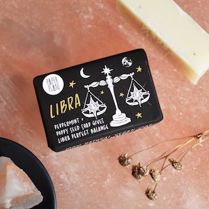 Puede incluir: Pastilla de jabón negra y dorada con un símbolo del zodiaco Libra y el texto "Peppermint + Poppy Seed Soap Gives Libra Perfect Balance".