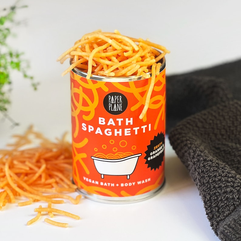 May include: A red and white can of bath spaghetti with the text "Paper Plane Bath Spaghetti" and "Zesty Orange Grapefruit Vegan Bath + Body Wash". The can is filled with orange bath spaghetti.