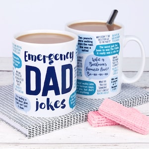 May include: White ceramic mug with a blue and white design. The mug features the text "Emergency Dad Jokes" and several humorous jokes in speech bubbles. The mug is filled with a brown liquid and a silver spoon is resting on the rim.