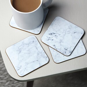 May include: Four white marble coasters with a gray veining pattern. The coasters are square and have a brown border.