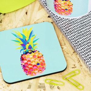 May include: A colorful geometric pineapple design on a light blue square coaster. The pineapple is made up of various colors, including yellow, orange, pink, blue, and green.