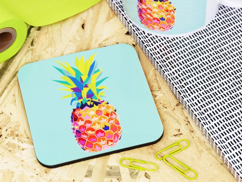 May include: A colorful geometric pineapple design on a light blue square coaster. The pineapple is made up of various colors, including yellow, orange, pink, blue, and green.