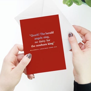 May include: A red Christmas card with a white envelope. The card features a humorous Christmas carol with the text "Quark! The herald angels sing, no dairy for the newborn king" and the caption "Millennial Christmas Carol #24".