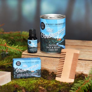 Op de afbeelding: Een natuurlijke gezichts- en baardverzorgingsset met de woorden "The Outdoorsman" op de verpakking. De set bevat een blikje met baardverzorgingsproducten, een zeep, een fles baardolie en een houten kam.
