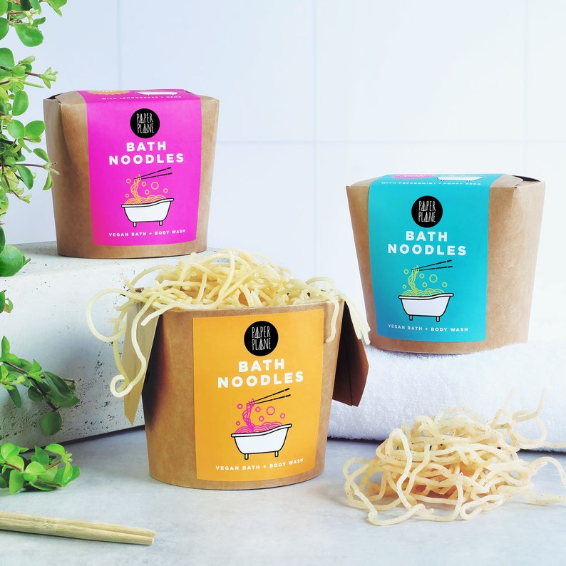 May include: Three brown cardboard containers with pink, blue, and yellow lids. Each container has a white label with the text "Paper Plane" and "Bath Noodles" in black. The containers are filled with yellow noodles. The containers are on a white surface.