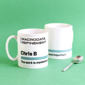 May include: Two white ceramic mugs with black text and blue stripes. One mug has a handle and reads "MACRODATA REFINEMENT", "Chris B", and "The work is mysterious". The other mug says "and important". A silver spoon is next to them.
