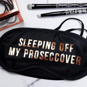 May include: Black sleep mask with the text "SLEEPING OFF MY PROSECCOVER" in gold lettering. The image also includes a rose gold framed box with brown hair ties and two black makeup brushes.