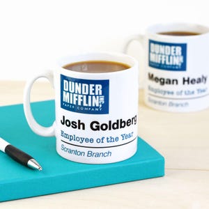 May include: Two white ceramic mugs with coffee. One mug features the "Dunder Mifflin" logo and the text "Josh Goldberg, Employee of the Year, Scranton Branch." The second mug has the same logo and "Megan Healy, Employee of the Year, Scranton Branch."