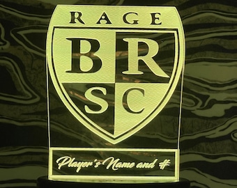 May include: A yellow and black acrylic night light with a shield-shaped design. The shield has the letters "BRSC" inside and the word "RAGE" above it. The text "Player's Name and #" is below the shield.