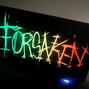 Może przedstawiać: Czarna prostokątna powierzchnia z napisem "FORSAKEN" w neonowym gradiencie zieleni, żółci i czerwieni. Tekst ma czcionkę grunge. Na dole widoczne jest małe niebieskie światło.