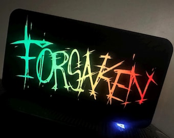 May include: A black rectangular surface with the word "FORSAKEN" in a neon gradient of green, yellow, and red. The text has a distressed, edgy font. The surface has a small blue light at the bottom.