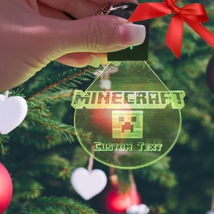 May include: A green and white Minecraft Christmas ornament with the word "Minecraft" and a pixelated creeper face. The ornament has a space for "Custom Text" and is hanging on a Christmas tree.