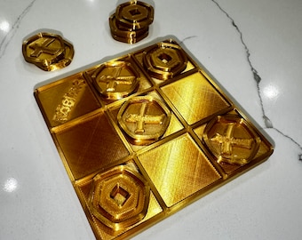 May include: A gold-colored 3D printed tic-tac-toe game board with nine squares. The board has a textured surface and the squares are outlined in black. There are four gold-colored game pieces with a cross design and one gold-colored game piece with a diamond design.