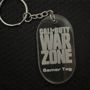 Może przedstawiać: Przezroczysty plastikowy brelok z wygrawerowanym napisem "Call of Duty War Zone Gamer Tag" w kolorze białym.
