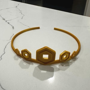 May include: A gold-coloured crown with a series of geometric shapes. The crown features a circular band with hexagonal and diamond-shaped accents. The central hexagon has a square cut-out. The crown is made of a solid material.