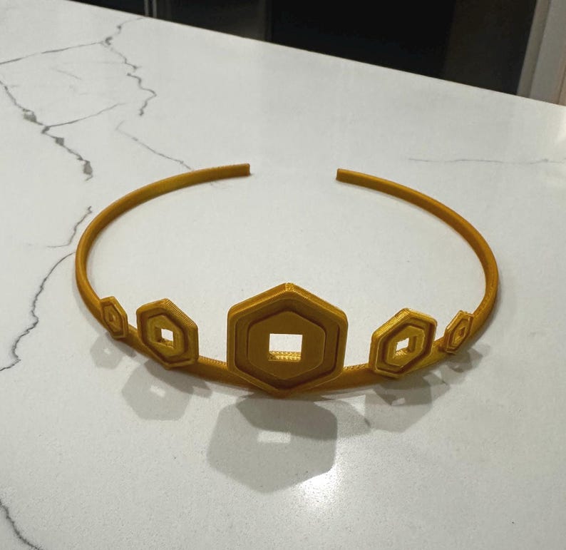 May include: A gold-coloured crown with a series of geometric shapes. The crown features a circular band with hexagonal and diamond-shaped accents. The central hexagon has a square cut-out. The crown is made of a solid material.