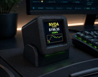 May include: A black, cube-shaped digital stock ticker device with a textured frame. The screen displays NVDA, NVIDIA, $198.35, +0.94%, and a green and red candlestick chart. A subtle green light glows from the base of the device on a desk.